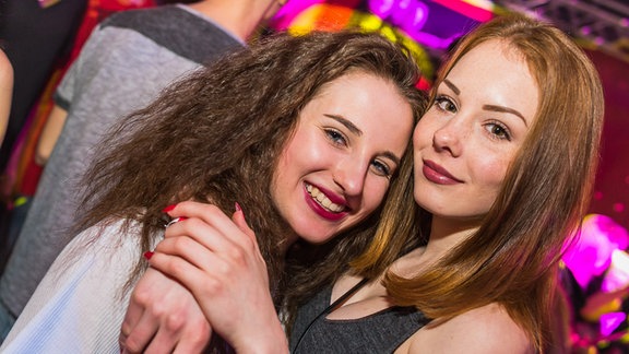 Zwei Mädchen schauen lachend in die Kamera. Sie befinden sich auf einer Party und umarmen sich.