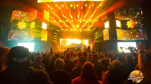 Eine Menschenmasse steht beim SonneMondSterne-Festival unter freiem Himmel vor einer großen Bühne mit orangeleuchtenden Bildschirmen.