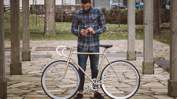 Ein junger Mann mit Bart im Holzfäller-Hemd schaut aufs Smartphone. Vor ihm steht ein Rennrad mit goldenem Rahmen und weißem Lenker.