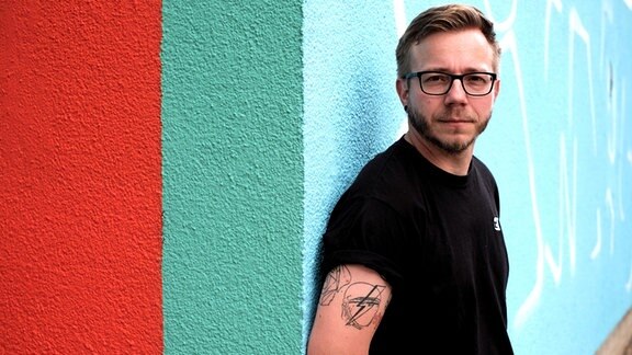 DJ Filburt steht angelehnt an eine bunte Wand. Er trägt eine Brille, schwarzes einfaches T-Shirt, blonde Haare. Auf dem rechten Oberarm hat er ein Tattoo.