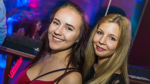Zwei junge Frauen lächeln in die Kamera, im Hintergrund tanzende Leute
