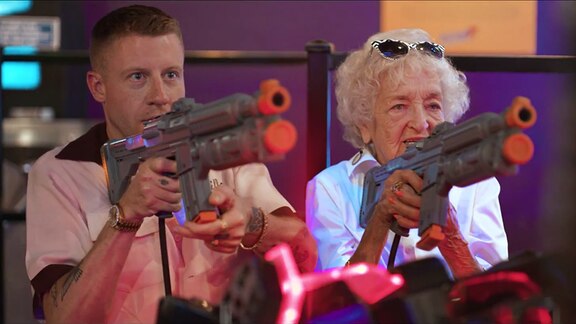 Zwei Menschen sitzen vor einem Spieleautomaten und zielen mit einer Spielzeugpistole