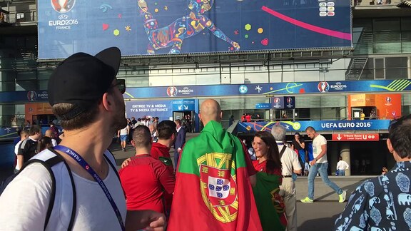 Mark Forster steht vor dem Stade de France in Paris. Er schaut über die Schulter zurück aufs Stadion und bewegt sich davon weg. Im Hintergrund sind portugiesische und französische Fans zu erkennen.