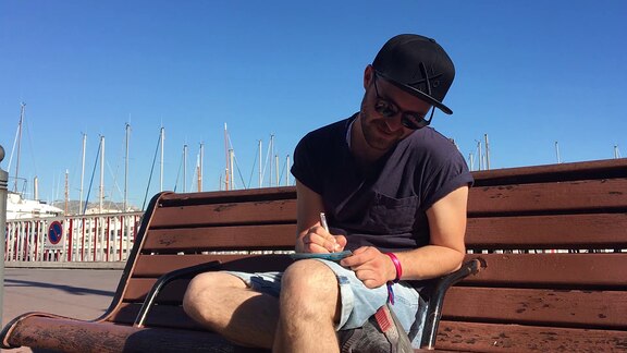 Mark Forster sitz auf einer Bank am Hafen von Marseille. Auf seinen überschlagenen Beinen liegt eine Postkarte, die er beschreibt.
