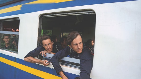 Die Brüder Hansen & Paul Hoepner schauen aus dem Fenster eines fahrenden Zuges. Ihre Haare wehen im Wind. Einer der beiden hält einen Selbstauslöser für die Fotokamera in der Hand. Im Hintergrund sind weitere Fahrgäste zu erkennen. Sie sind asiatisch.