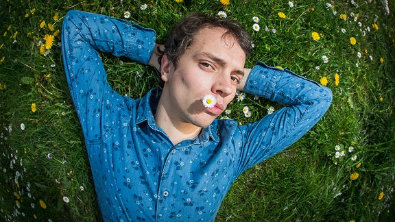 Frans Zimmer (Alle Farben) liegt in blauem Hemd auf einer sattgrünen Sommerwiese und hat  'ne Gänseblümchen im Mund. Er die Arme hinter dem Kopf verschränkt und schaut in die Kamera. 