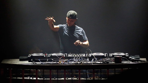 Avicii steht während eines Gigs hinter einem mächtige DJ Pult. Er trägt ein graues T-Shirt und ein Basecap mit nach hinten gedrehtem Schild. Seine rechte Hand ist etwa bis zur Waagerechten erhoben. Das ganze Foto besteht aus eher düsteren Grautönen.