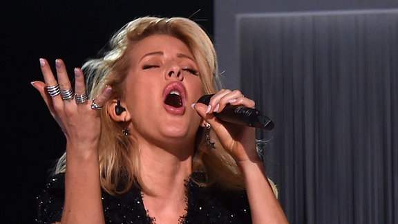 Ellie Goulding ist mit geschlossenen Augen während sie einen Song performt zu sehen. Auffällig sind die 4 silbernen Ringe an ihrer rechten Hand.