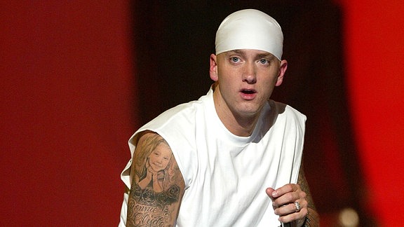 Eminem während eines Konzerts im Jahr 2004 in Culver City/Californien. Er tägt ein ärmelloses weißes Shirt und weißes Kopftuch. Seine Tattoos des rechten Arms sind gut zu sehen. Das Mikro hält Eminem in der linken Hand, etwa in Brusthöhe und blickt links an der Kamera des Fotografen vorbei.