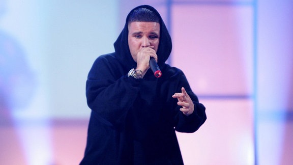 Der Rapper Fler trägt einen schwarzen Kapuzen-Pullover und spricht in ein Mikro. Er steht auf einer Bühne, hinter ihm leuchten lila-blaue Lichtstrahler.