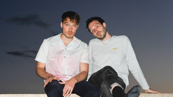 Zwei Männer in weißen Hemden und dunklen Hosen sitzen entspannt nebeneinander.