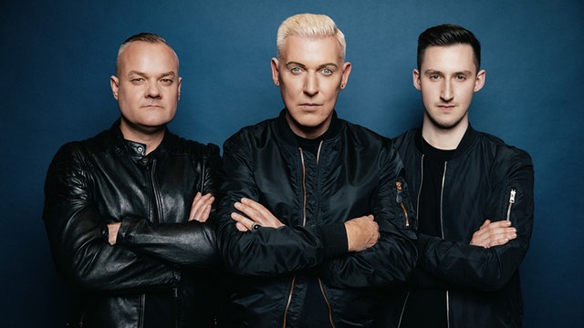 Techno-Band Scooter, die drei Mitglieder schauen mit verschränkten Armen in die Kamera, dunkelblauer Hintergrund