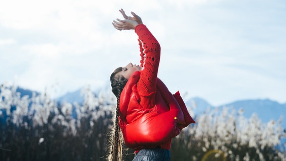 Musikerin CARY vor bergiger Kulisse in glänzender roter Puffer-Jacke und bläulich-schimmernder Hose mit  Kopf und Armen den Himmel zugewandt