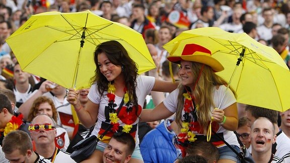 Public Viewing, zwei Mädchen sitzen auf Schultern mit gelben Schirmen in einer Menschenmenge mit deutschen Fußballtrikots