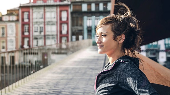 Ein Mädchen mit hochestecktem Haar steht angelehnt auf einer Brücke und schaut entspannt und zuversichtlich in die Ferne. Im Hintergrund sind Häuserfassaden zu erkennen. Das Bild scheint in Südeuropa aufgenommen zu sein.