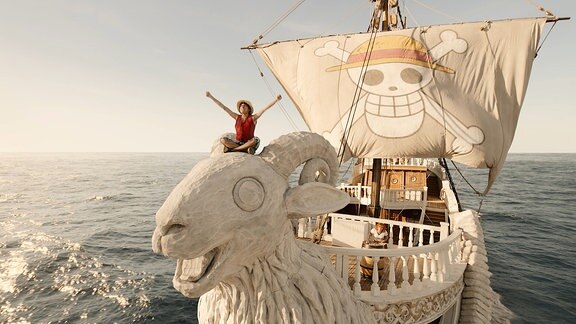 Hauptcharakter Ruffy steht am Bug eines weißen Schiffes, das vorn wie ein großer Ziegenkopf geformt ist. Hinter ihm weht die Piratenflagge. luffy streckt seine Arme gen Himmel.