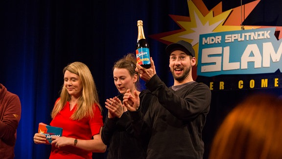 Beim SPUTNIK Slamedy hält Tobias Kunze eine Sektflasche mit der Aufschrift "Slamedy Winner" hoch.